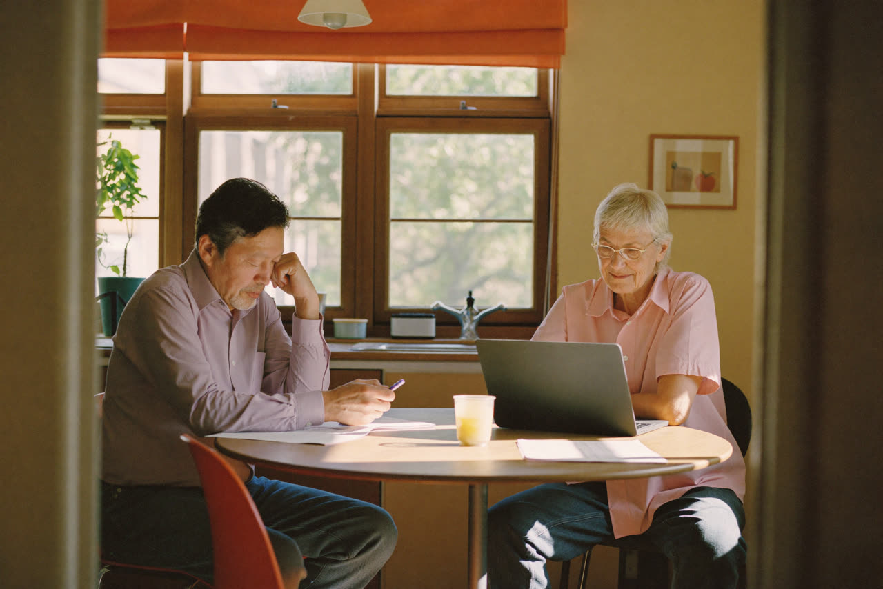 A couple at a sunlit kitchen table with a laptop and paperwork